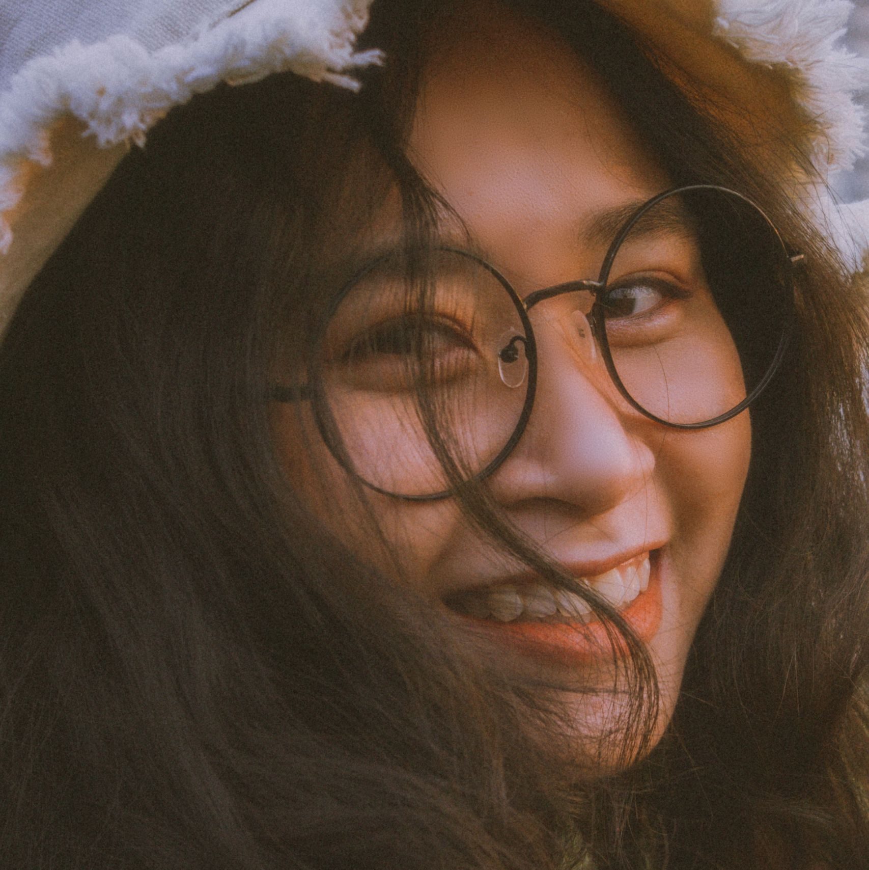 Portrait of a smiling woman wearing glasses and hat, with long hair blown by the wind.
